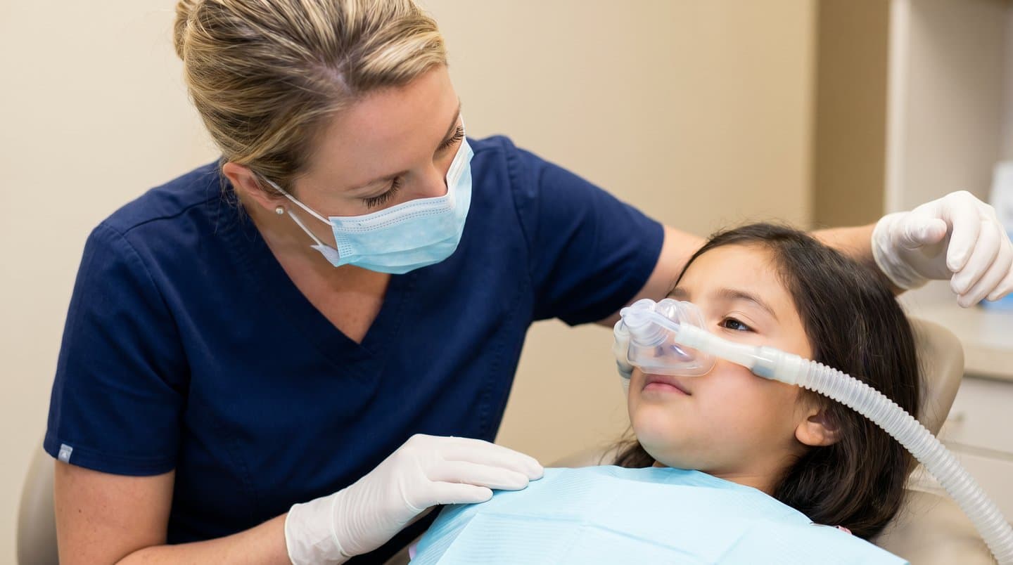 A relaxed child in a dental chair wearing a nitrous oxide nose mask while a dental professional in scrubs provides care